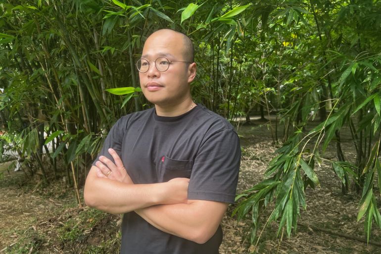 A portrait of Sebastien Lai. He is wearing a black t-shirt and standing with his arms crossed in a park