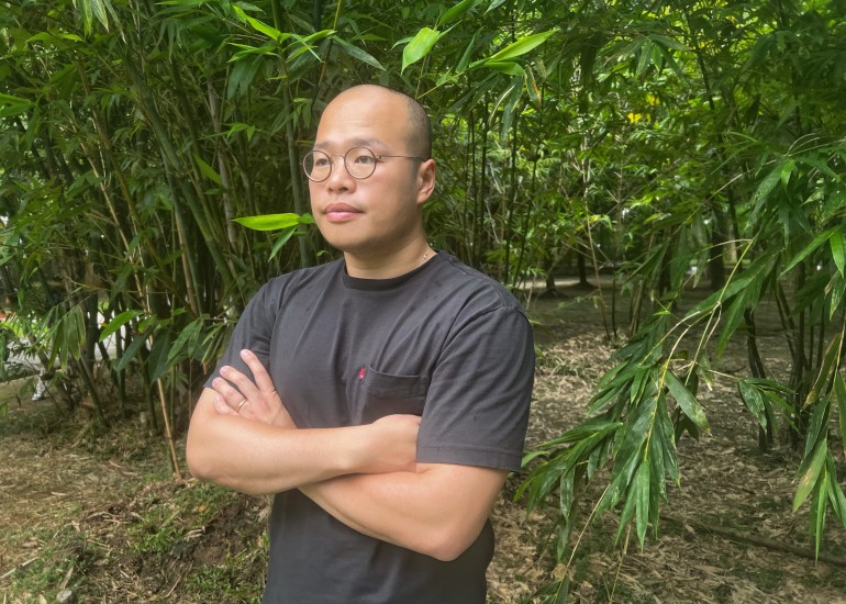 A portrait of Sebastien Lai. He is wearing a black t-shirt and standing with his arms crossed in a park
