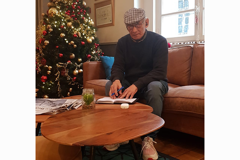 A photo of a man sitting on a sofa and writing something in a notebook on top of the table in front of him with a Christmas tree in the corner of the room next to him.