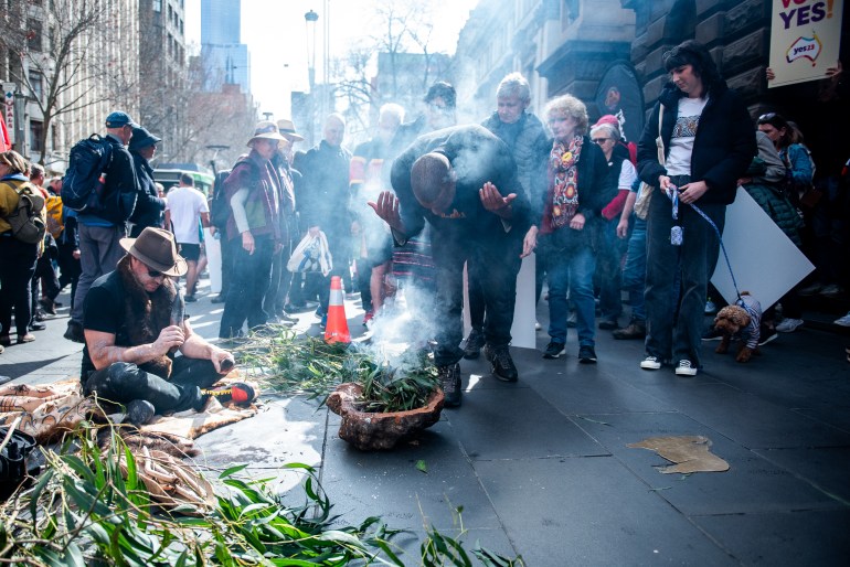 A man conducting a traditional smoking ceremony on the road outside Melbourne Town Hall. He is leaning over a wooden bowl containing green leaves that are giving off smoke. A man to his side is speaking into a microphone. There are people watching the ceremony. 