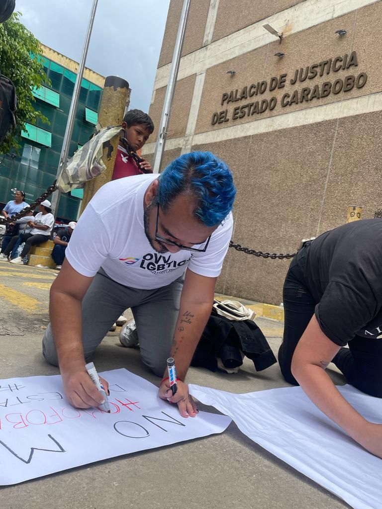 A man with blue hair kneels on the sidewalk to write a protest message on poster board outside the Carabobo courthouse in Venezuela.