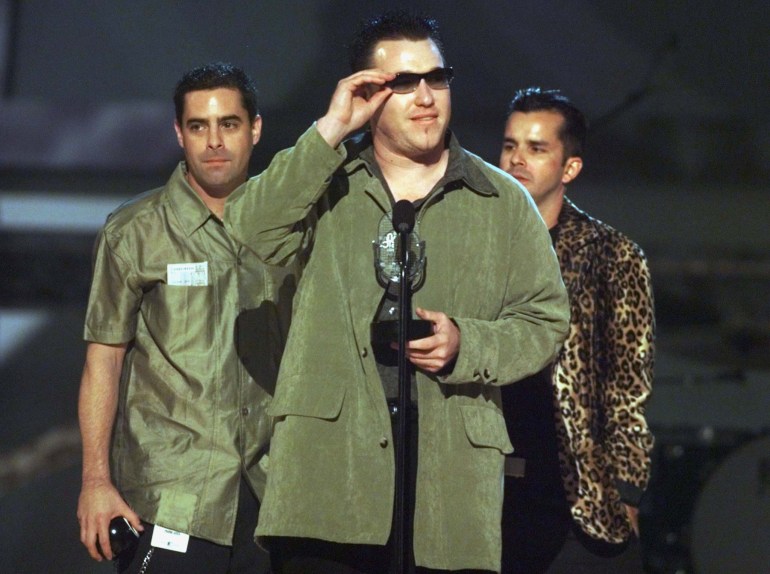 A man in an olive green coat stands in front of a microphone on stage, wearing sunglasses. Two other men stand behind him. This photo depicts Steve Harwell at the WB Radio Music Awards show at the Mandalay Bay Events Center in Las Vegas on October 28, 1999.