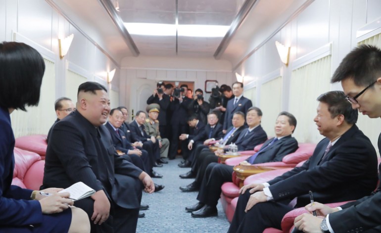 Kim holding discussions with Chinese officials on board his private train. The walls are white and pink armchairs face each other down either side of the carriage. The carriage curtains are closed.