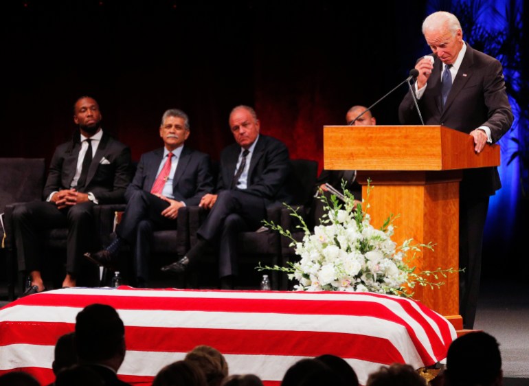 Joe Biden stands at a podium dabbing his eyes, as a casket covered with an American flag sits in front of him.