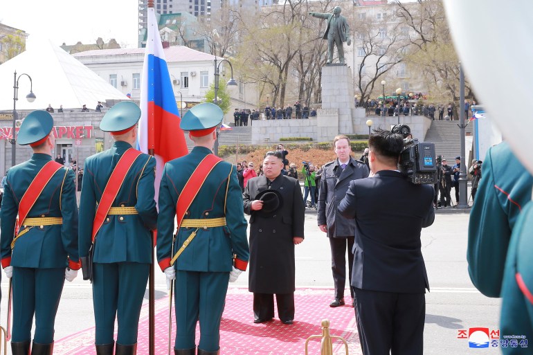 Kim Jong Un stands on a red carpet, surrounded by soldiers and officials.