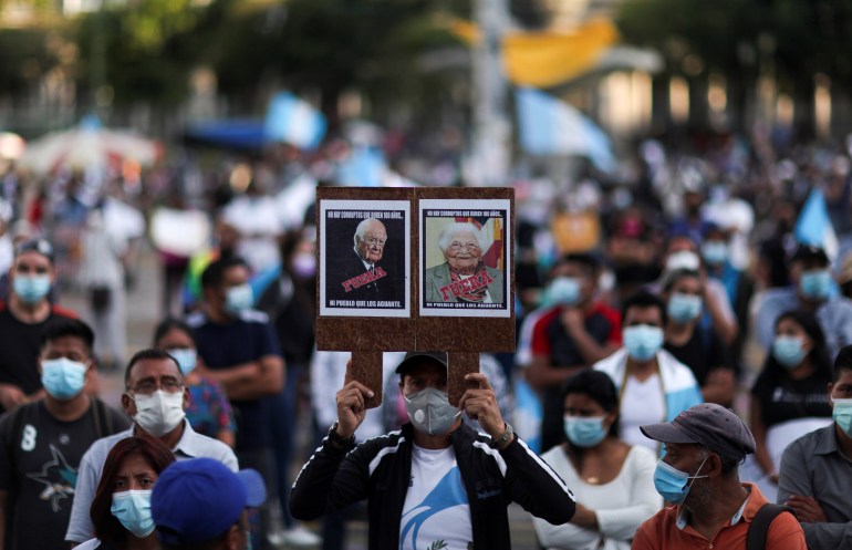 A protester, dressed in the colors of Guatemala's flag, holds up twin portraits of the president and the attorney general that have been digitally aged. Across each one is written the word "fuera" or "out".