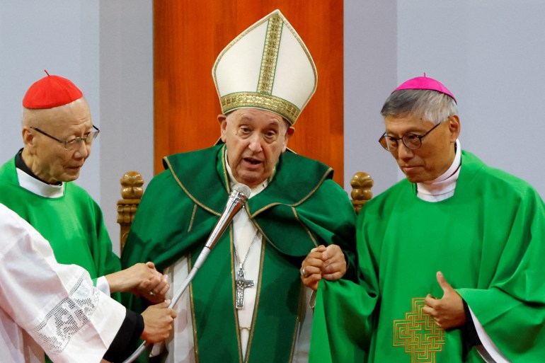 The pope supported by ardinal John Tong Hon and Hong Kong's Archbishop Stephen Chow. He is wearing green robes and a white mitre with gold trimmings. speaking into a microphone