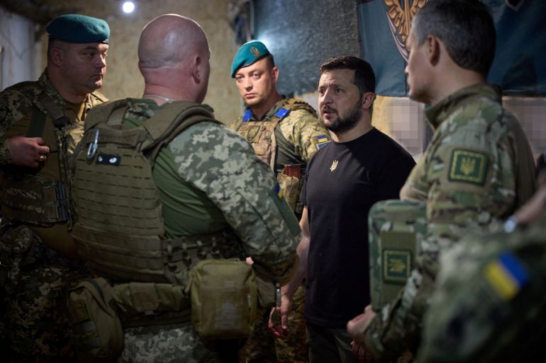 Ukrainian President Volodmyr Zelenskyy visiting frontline troops in the eastern Donetsk region. He is wearing a black T-shirt. The soldiers around him are in uniform. They look serious.