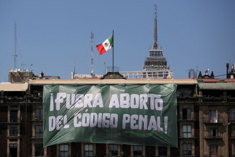 A green banner with white text hangs from a building topped by a Mexican flag.