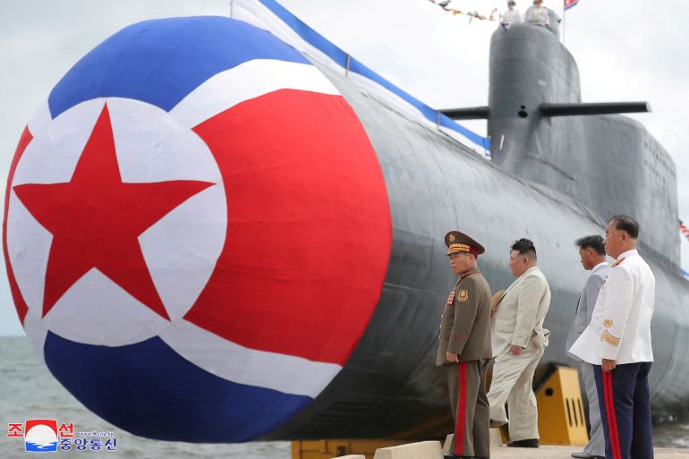 Il nuovo sottomarino della Corea del Nord coperto dalla bandiera nordcoreana mentre scivola in acqua.  Kim Jong Un sta assistendo al lancio accompagnato da alcuni ufficiali militari.
