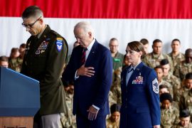 President Joe Biden, standing between two service members, bows his head and puts his hand over his heart. Behind him are people in military fatigues, standing against a gigantic US flag.