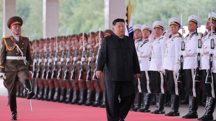 Kim Jong Un walking along a red carpet in front of a line of soldiers standing to attention. He is accompanied by another soldier who is goose stepping beside him