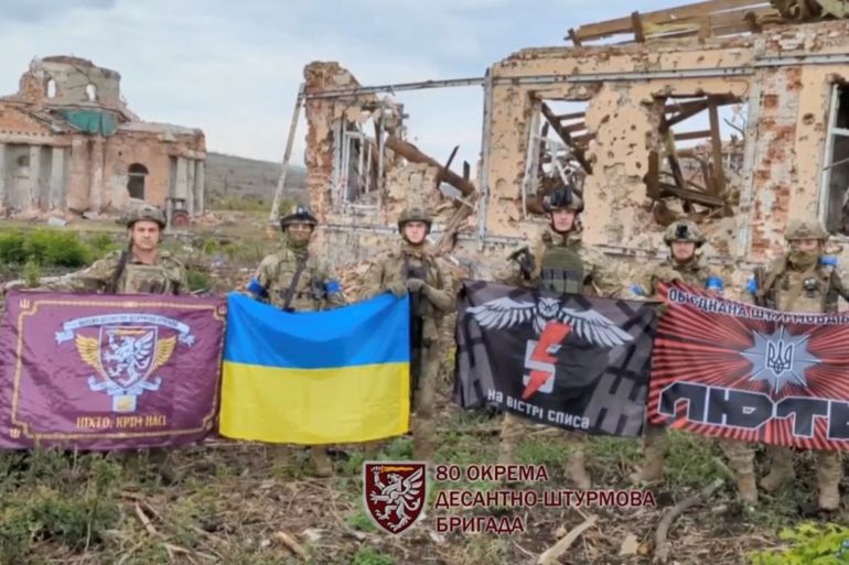 Soldiers hold the Ukraine flag and flags from their fighting groups in front of ruined buildings in Klishchiivka