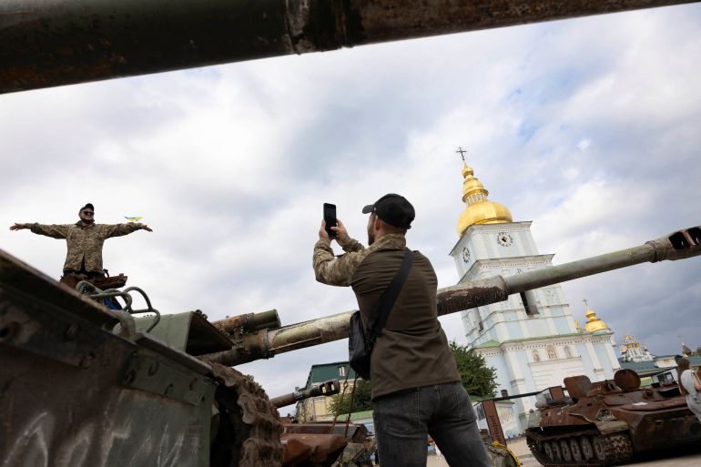 A man stands on a captured Russian tank in Kyiv. He has his arms outstretched and a friend is taking his picture.