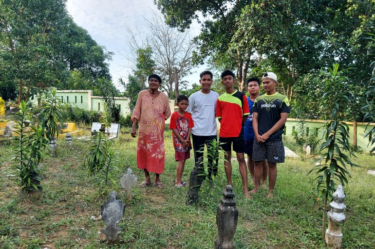 Halimah standing with her grandchildren in the cemetery in Sembulang. She is wearing a flowery peach coloured dress and looks content. She has five grandsons standing next to her. Carved stones mark each grave and there are small bushes