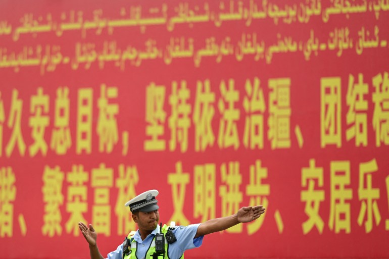 Um policial orientando o trânsito em Xinjiang.  Um outdoor vermelho atrás dele proclama a necessidade de manter o Estado de direito na região.  As palavras são escritas em amarelo na escrita uigur e em caracteres chineses.