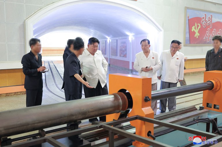 Kim Jong Un inspecting a munitions factory. He is standing next to some steel tubes. An official is explaining something to him.