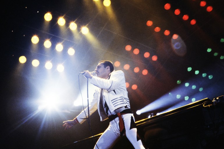 Freddie Mercury perforning on stage in 1984. He's wearing tight white trousers with black markings on the side and a white studded jacket