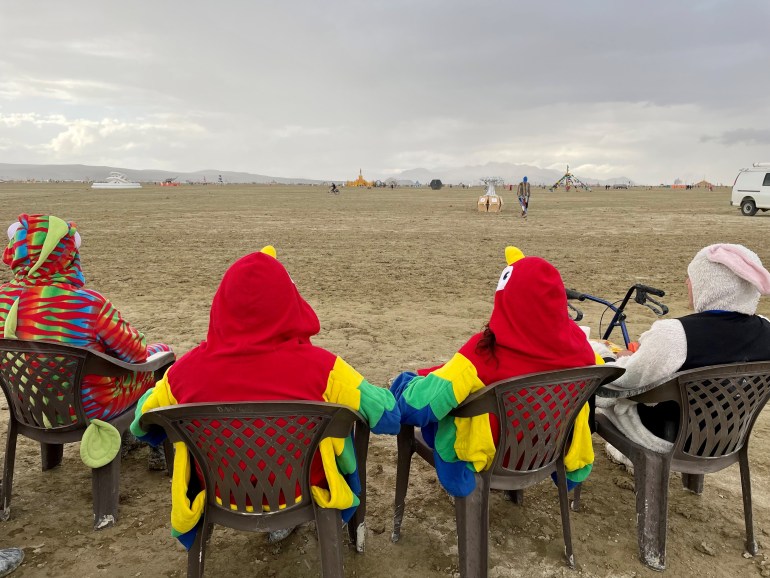 Festival goers sitting in camp chairs looking out at the Burning Man site. They are dressed in red, green and yellow onesies.