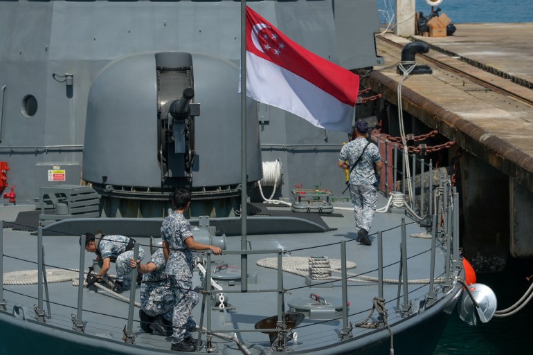 A close up of a Singapore navy ship.. The Singapore flag is flying. There are sailors working on deck