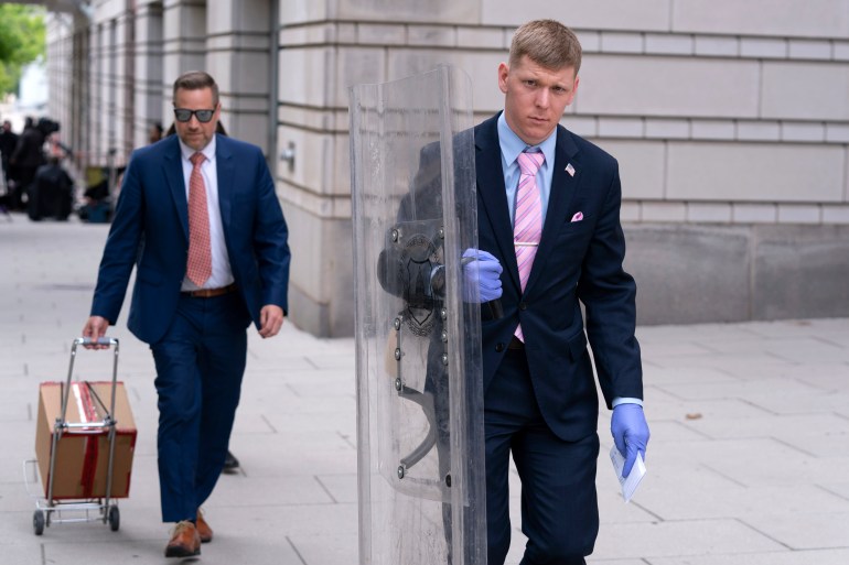 A man wearing blue evidence gloves and a suit and pink tie walks outside a courthouse with a clear plastic riot shield as evidence.