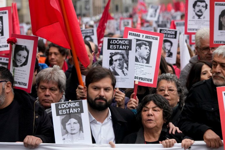 President Gabriel BOric stands amid a sea of protesters who wave red flags and hold black-and-white print-out photos of the people who went missing under Pinochet, labelled "Donde Estan?" Or "Where Are They?"