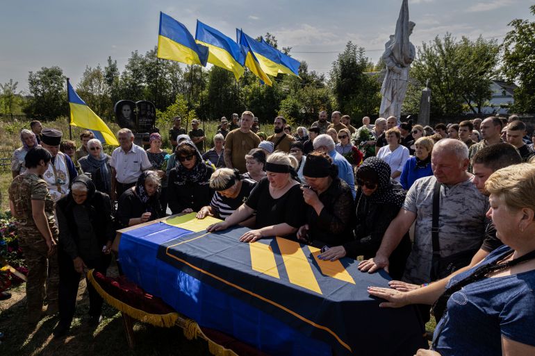 A family mourns their son, killed fighting for Ukraine near Bakhmut. His mother and other family members have their hands on the coffin, which is draped in blue and yellow. Ukrainian flags are flying behind them.