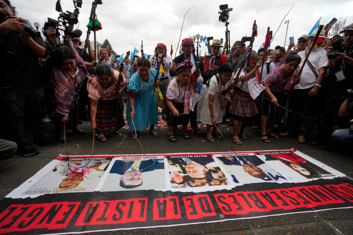 Indigenous women use sticks to hit a line of photos laid out on the ground, representing key figures in Guatemala's government.