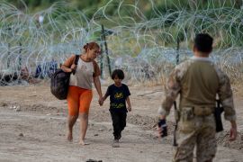 Migrants who crossed into the U.S. from Mexico walk past concertina wire lining the banks of the Rio Grande as they move to an area for processing, Sept. 21, 2023, in Eagle Pass, Texas. The woman holds the young child's hands, while a guard, facing away from the camera, looks on.