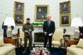 US President Joe Biden, in a suit, stands next to Ukraine President Volodymyr Zelenskyy, wearing military attire, in the Oval Office, where they stand in front of two beige chairs, a mantlepiece and a wall full of photos.