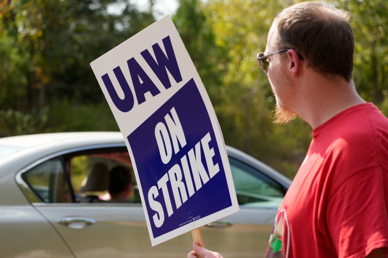 A man in a red shirt holds a picket sign that reads, "UAW On Strike," as a car passes him by.