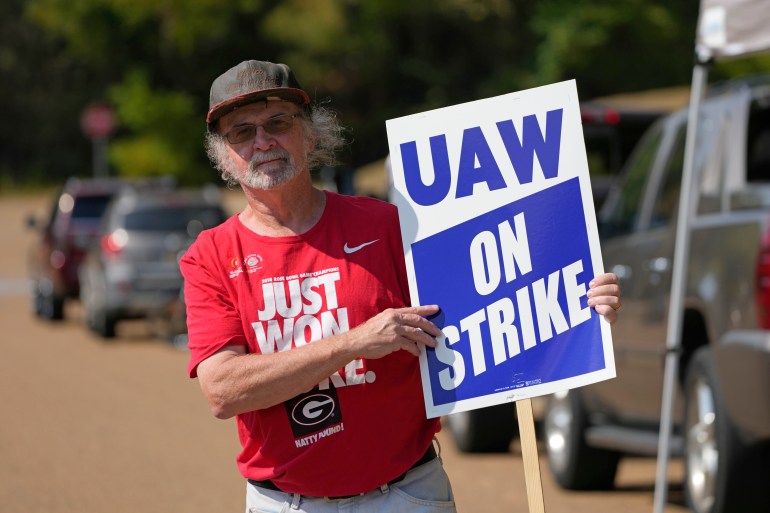 United Auto Workers member Ron Oglesby holds a picket sign outside the General Motors Customer Care and After-Sales facility in Brandon, Miss., Friday, Sept. 22, 2023. Unionized workers joined others in new nationwide walkouts as the labor standoff continues. Oglesby wears a hat and a red shirt, and he points to his picket sign that reads, "UAW on strike."