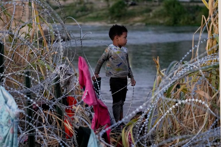 Migrants waits to climb over concertina wire after he and others crossed the Rio Grande and entered the U.S. from Mexico, Saturday, Sept. 23, 2023, in Eagle Pass, Texas. In the center of the photo stands a child, perched between the razor wire and the river. Shredded clothes hand from the razor wire.