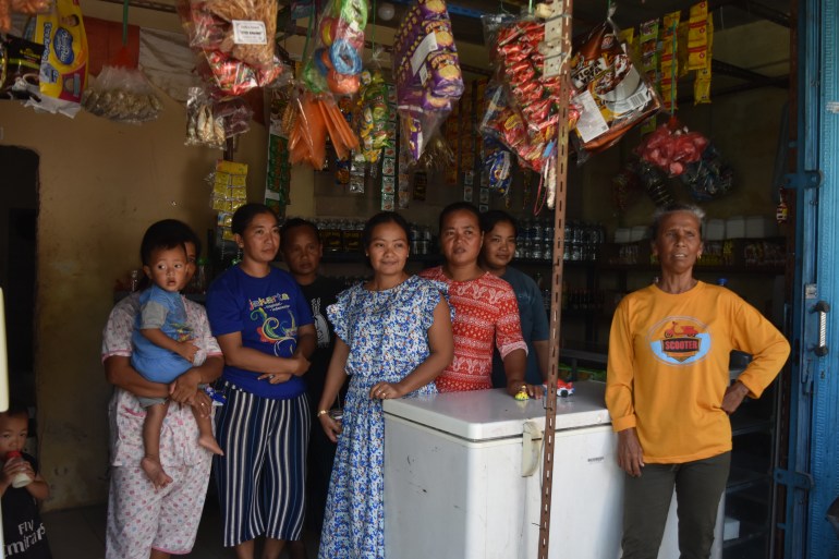 Nor (far right) standing in a sundry shop in Rempang. She is standing near a large chest freezer and there are products hanging from the ceiling. Other women are standing the other side of the freezer