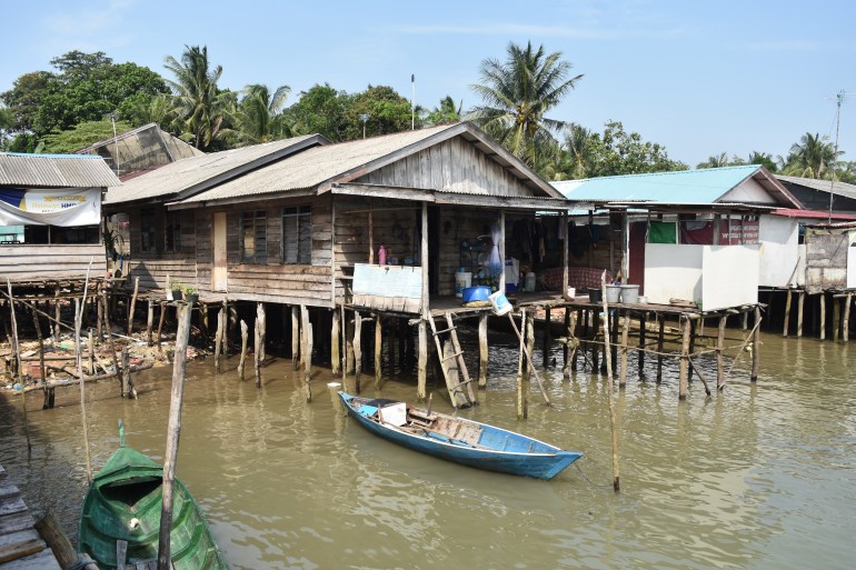 A view of wooden houses built on stilts at the water's edge. Narrow wooden boats are tied up outside some of the houses. There are palm trees behind.