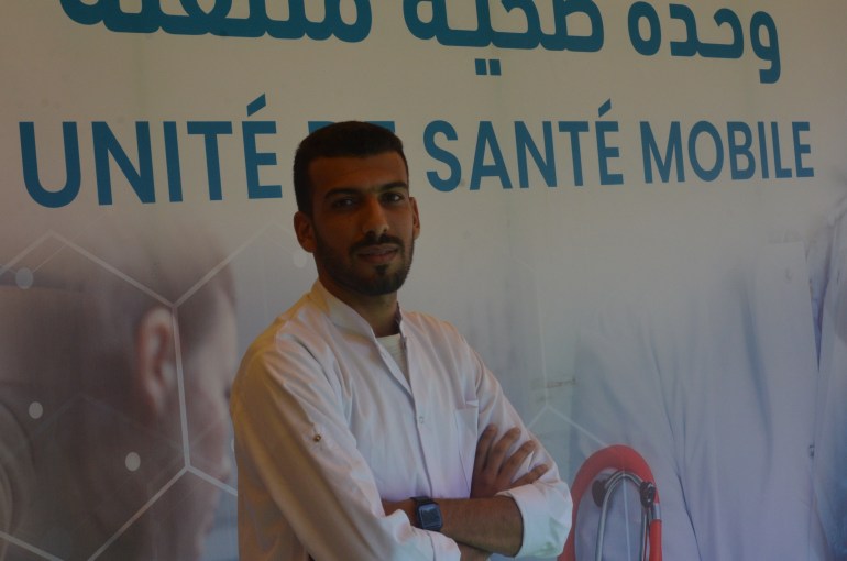 A doctor, dressed in a white coat, stands against a sign that reads: "Unité Santé Mobile."