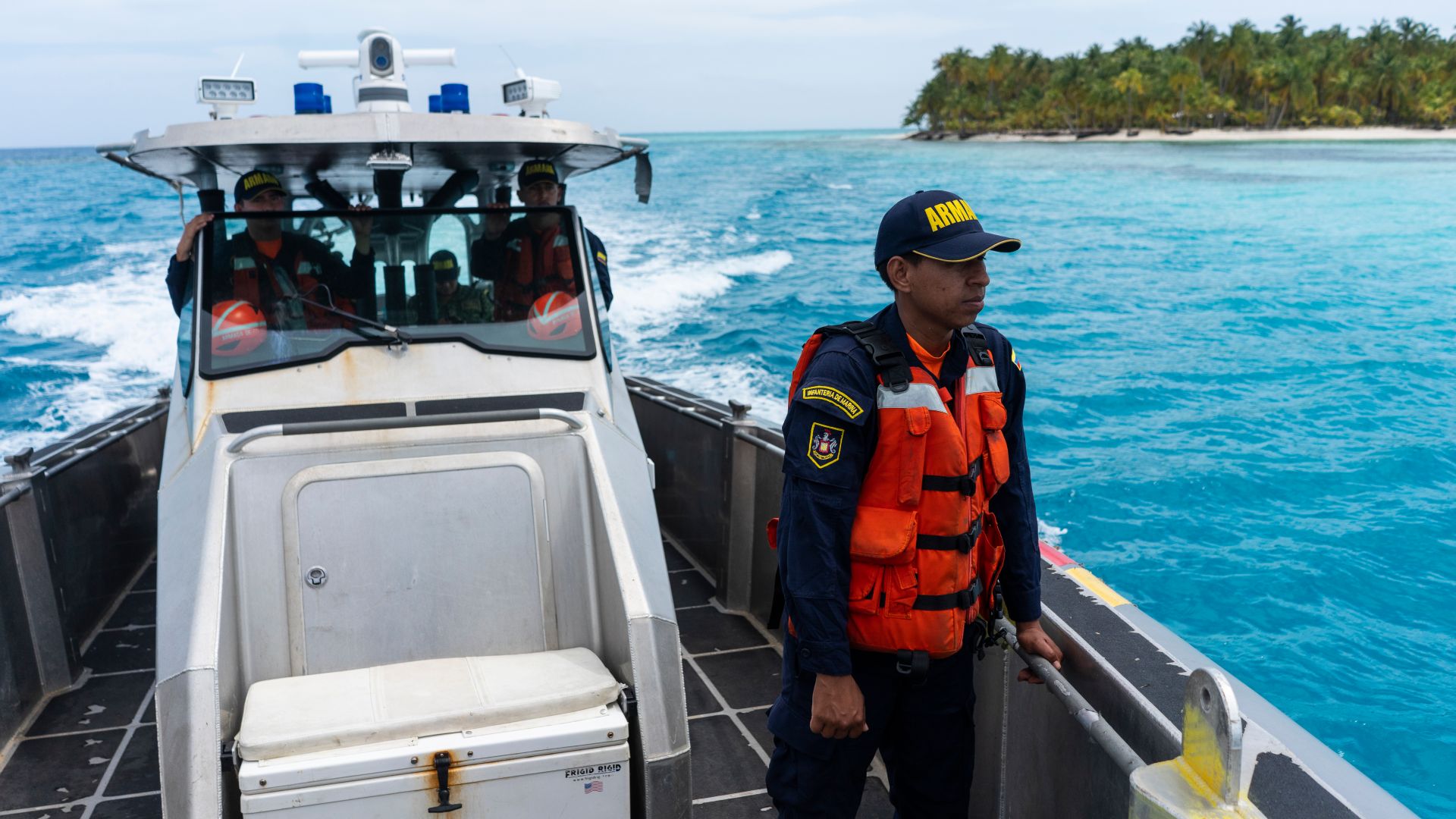 A Navy official, dressed in a ballcap, a long-sleeved uniform and an orange life vest stands aboard a boat that plies its way through the blue Caribbean waters.