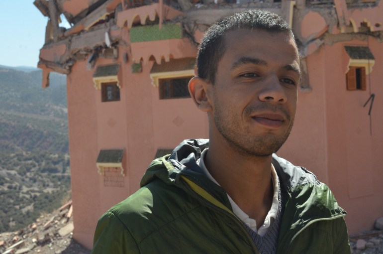 A young man in a green hoodie stands in front of a Moroccan town