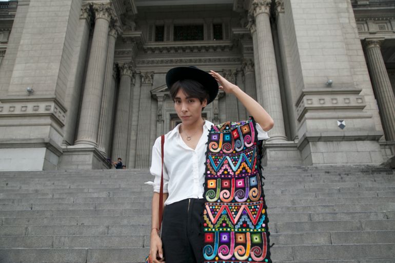 A young Peruvian man lifts one arm to adjust his dark hat. He wears a crisp white shirt, and over his shoulder is an Indigenous belt, colorfully embroidered. He stands on the steps of Peru's Supreme Court, an imposing neoclassical building.