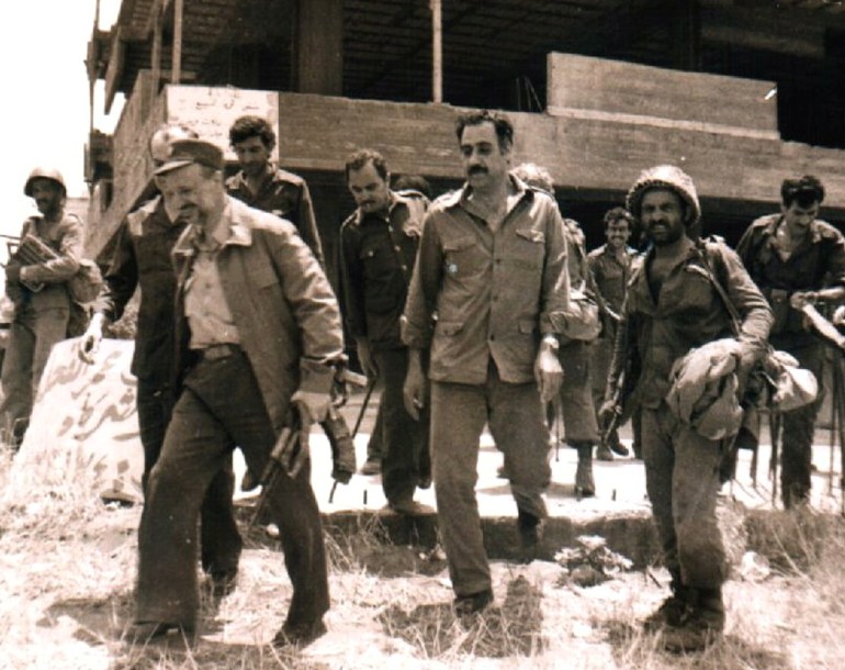 Yasser Arafat walks with a group of people in military fatigues and helmets in an undated, sepia-coloured photo.