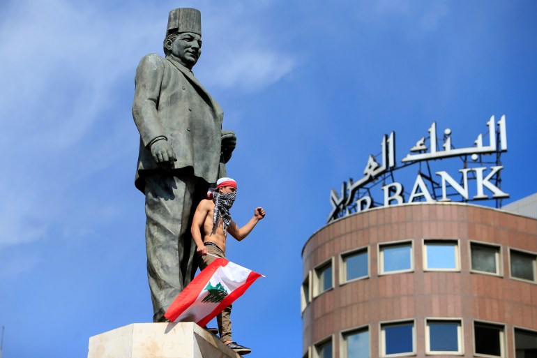 A protester, shirtless and carrying a Lebanese flag, stands at the base of a large statue. The sign for a bank looms from a nearby building.