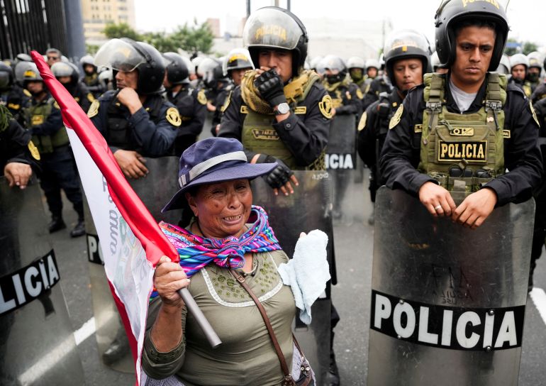 An Indigenous woman walks in front of a group of Peruvian riot police on the street, as she carries a Peruvian flag in protest.