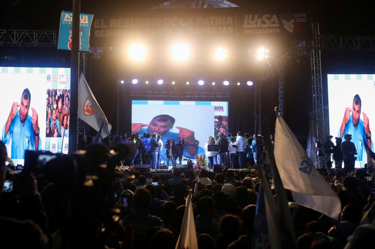 Ex-president Rafael Correa appears on large screens in front of a dark but crowded room, where some supporters wave flags.