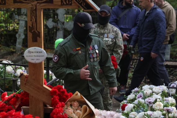Wagner fighters at Prigozhin's grave. They are in fatigues and have covered their mouths with black masks. There are flowers around the grave.