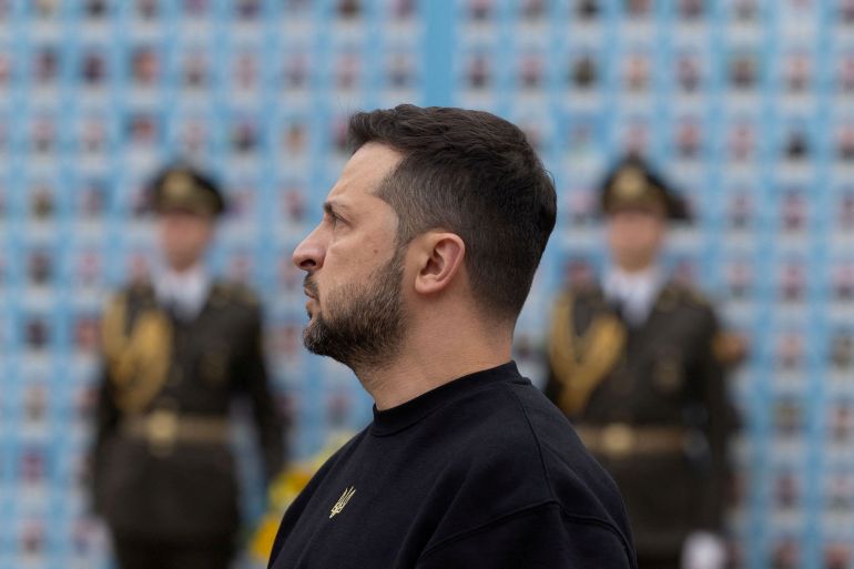 Volodymyr Zelenskyy pays his respects at the emory Wall of Fallen Defenders of Ukraine. He is photographed in profile. There are two soldiers in ceremonial dress in the background.