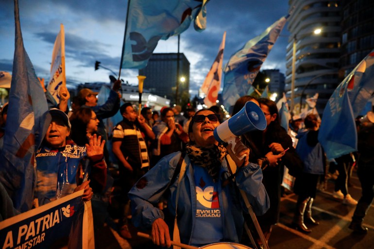 Supporters of Ecuadorian presidential candidate Luisa Gonzalez rally outside at twilight, waving flags, carrying banners and wearing blue campaign shirts.