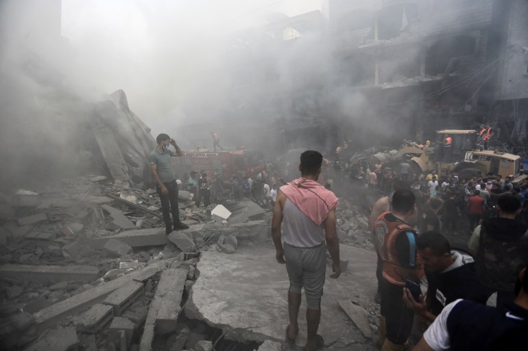 A man with a rose-coloured cloth around his shoulders stands amid the rubble of a bombed refugee camp, as dust hangs in the air. Others look on amid the smoke.