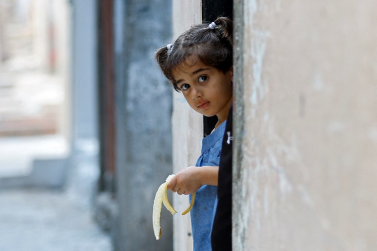 A little girl with pigtails holds an empty banana peel as she peers out from the door of a concrete house.