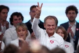 a man in a white shirt with a red heart sticker on his chest holds a peace sign with his fingers with supporters behind him