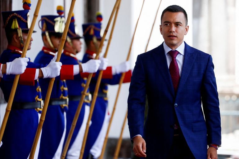 Daniel Noboa, wearing a blue suit and red tie, walks past a line of ceremonial guards, holding what appear to be staffs.